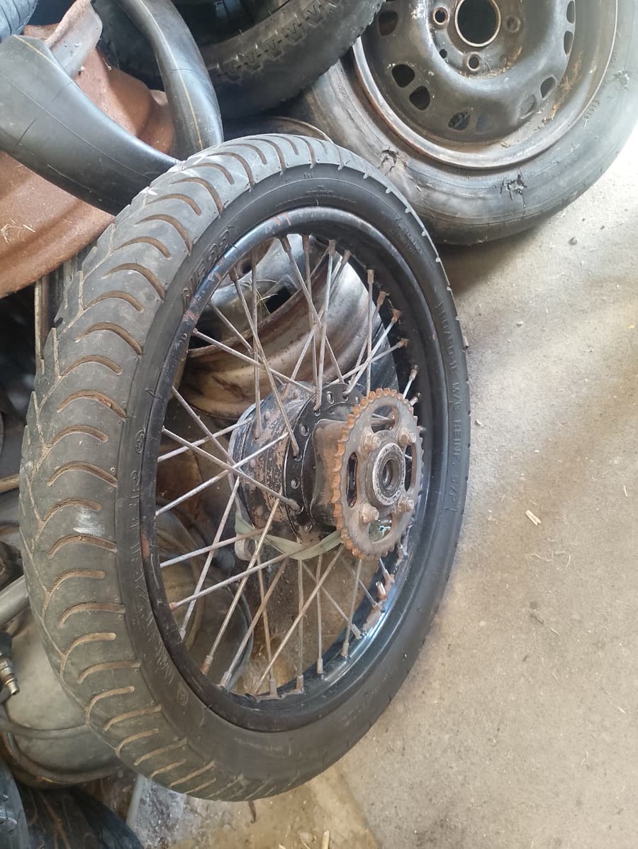 Una rueda de moto con rayos y corona, apoyada entre cubiertas en el taller